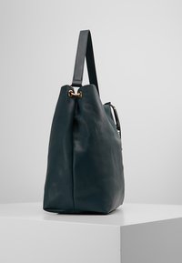 Tyrkysově modrá kožená kabelka s hrubou strukturou, volným tvarem, dlouhým ramenním popruhem a zlatými kovovými detaily na horní straně.