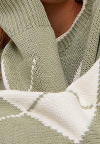 Pull en tricot vert clair à col en V, avec un motif en losanges blancs le long de l'ourlet et des poignets. La texture est douce et agréable au toucher.