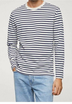 Homme portant une chemise à manches longues blanche avec des rayures horizontales bleu marine et un jean bleu clair, main droite dans la poche, sur un fond uni.