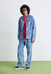 Chaqueta y pantalones de mezclilla con tela de color azul claro y estampados gráficos en blanco, combinados con una camisa roja y zapatillas negras.