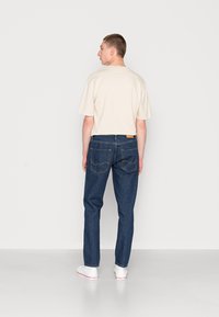 Camicetta beige a maniche corte abbinata a jeans di denim scuro con tasche posteriori e una patch in pelle color cuoio. Sneakers bianche completano l'outfit.