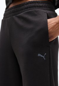 Κοντινό πλάνο μαύρου αθλητικού παντελονιού με ελαστική μέση και κεντημένο λογότυπο Puma κοντά στην τσέπη πλαϊνής πλευράς, με το χέρι μέσα στην τσέπη.