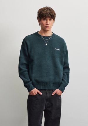 Jeune homme aux cheveux bruns portant un pull vert foncé avec un logo blanc et un jean noir, debout les mains dans les poches devant un fond clair.