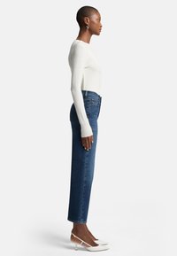 Top bianco a maniche lunghe, jeans blu a vita alta, vista di profilo, silhouette aderente, tessuto liscio, tacchi bianchi appuntiti con tacco basso.