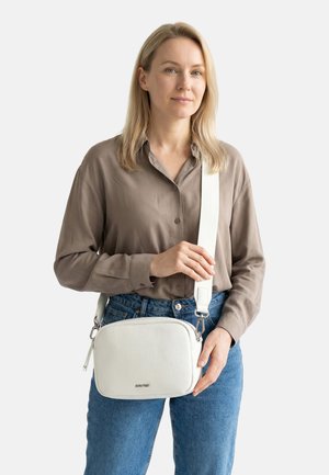Frau mit blondem Haar, die ein braunes Hemd und blaue Jeans trägt, hält vor einem weißen Hintergrund eine weiße Leder-Umhängetasche mit breitem Gurt.
