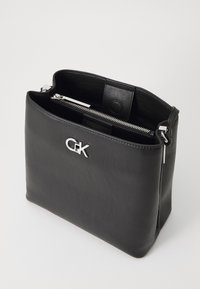 Borsa a tracolla in pelle nera con logo argentato, chiusura con zip e scomparti interni. Texture liscia, forma rettangolare e design minimalista.