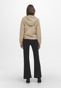 Beżowa kurtka z kapturem, z elastycznymi detalami przy końcach i mankietach, w połączeniu z czarnymi spodniami dzwonami i wysokimi obcasami, pokazana od tyłu.