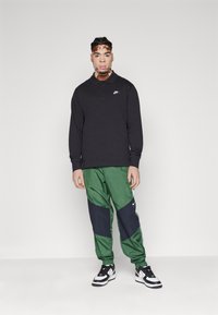 Polo preto de mangas longas com colarinho, calças desportivas verdes com painéis em azul-marinho e ténis em preto e branco com um design retro.