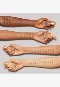 Quatre bras avec des teintes de peau différentes affichant des échantillons de fond de teint. Chaque bras tient une bouteille ; les nuances sont étiquetées à côté des échantillons pour identification.