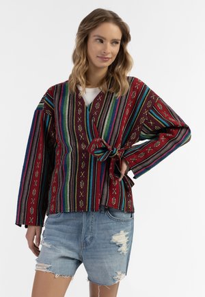 Rode en meerkleurige gestreepte wikkeljas met een strik in de taille, voorzien van een gestructureerde stof en decoratieve patronen. Draagt over denim shorts.