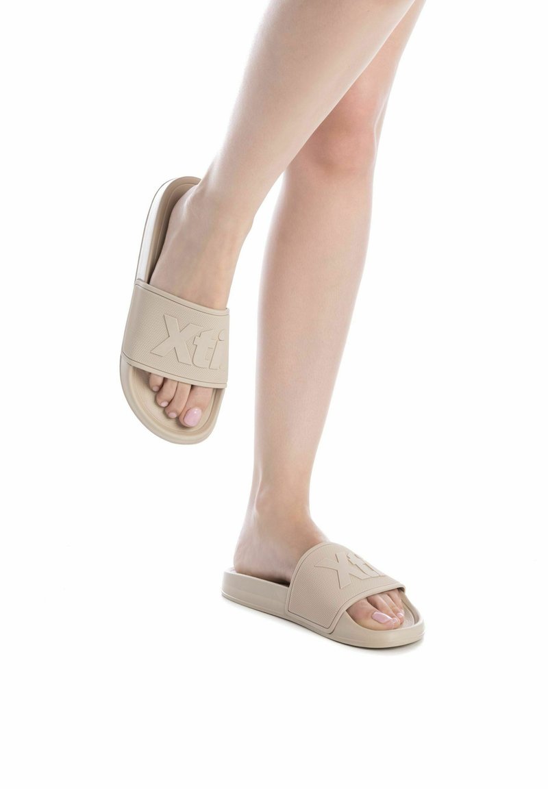 Chanclas de goma beige con una amplia correa que presenta el logotipo "Xti" en relieve, un diseño de punta abierta y una suela plana.