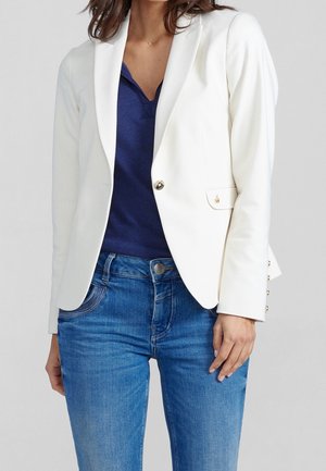 Frau trägt weißen Ein-Knopf-Blazer über marineblauem Hemd und blauen Jeans, steht mit der rechten Hand entspannt an der Seite.