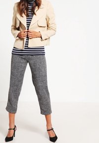 Giacca corta beige con spalle strutturate, top a righe blu e bianche, pantaloni cropped grigi con texture, e décolleté neri con cinturino alla caviglia.