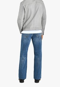Persona che indossa una giacca di pelle grigia chiara, una camicia bianca, jeans blu a gamba dritta e scarpe nere, vista di spalle contro uno sfondo bianco.