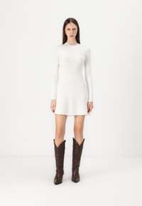 Robe longue blanche à manches longues avec une silhouette près du corps et une jupe évasée. Bottes de cowboy marron hauteur genou avec des détails de couture complexes.