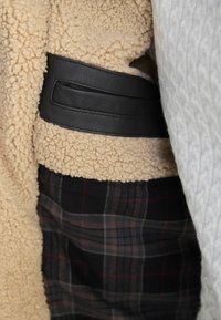 Beige textured jacket with black leather belt detail over dark plaid pants and a gray knit sleeve visible on the side.