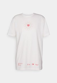 Valkoinen puuvillapaita lyhyillä hihoilla, jossa on keskellä punainen graafinen kuvio ja alareunassa teksti punaisella. Teksti sisältää sanat "Päättäväisyys Yli Negatiivisuuden."