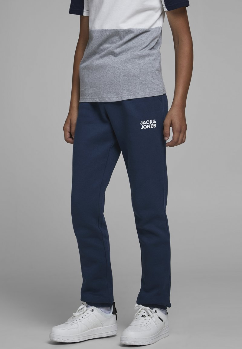 Pantaloni tuta blu navy con cintura elasticizzata, tasche laterali e logo bianco "JACK & JONES" sulla coscia. Abbinati a una maglietta grigia e bianca a blocchi di colore.