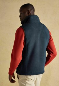 Tumši zilas vilnas veste ar augsto apkakli un reljefā "JOULES" logo, uzvilkta virs sarkanas garām piedurknēm krekla, piegriezuma forma pielāgota, sānu kabatas redzamas.