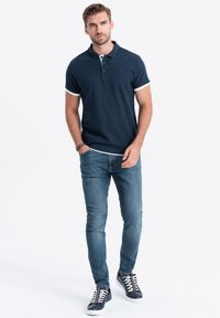 Námořnická modrá polo tričko s bílým límcem a akcenty na rukávech, v kombinaci se světle modrými džíny a námořnickými teniskami s bílou podrážkou a oranžovými detaily.