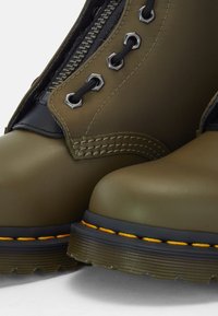 Gros plan sur des bottes en cuir vert olive avec des lacets noirs, des œillets métalliques, des semelles noires et des coutures jaunes sur un fond blanc.
