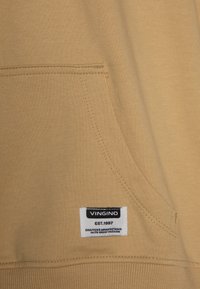 Felpa color beige con tasca a marsupio, orlo a costine e un'etichetta con logo in bianco e nero. Tessuto morbido con una superficie liscia.