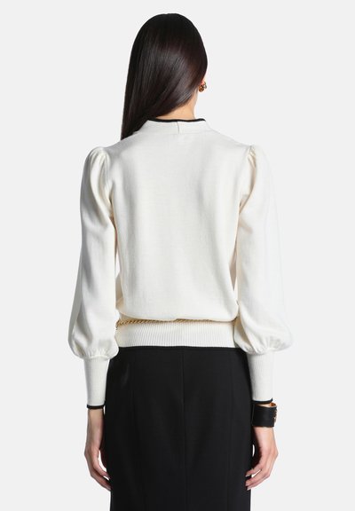 Femme aux longs cheveux foncés portant un pull blanc à manches bouffantes et une jupe noire, tournée de dos sur un fond clair uni.