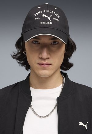 Młoda osoba nosząca czarną czapkę Puma, czarną kurtkę z logo Puma, białą koszulę i srebrny łańcuszek na szarym tle.