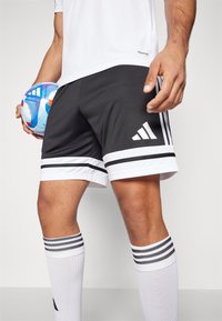 Mustat urheilushortsit, joissa on valkoiset raidat helman kohdalla, valkoisia teksturoituja koristeita ja kolme valkoista Adidas-raitaa sivussa, yhdistettynä valkoisiin sukkiin.