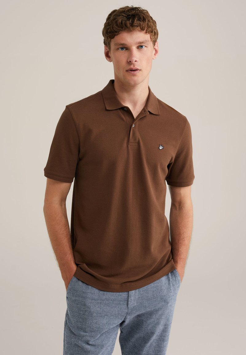 Camisa polo marrón de manga corta con cuello, que cuenta con un pequeño logo bordado en el lado izquierdo. La tela tiene una textura suave.