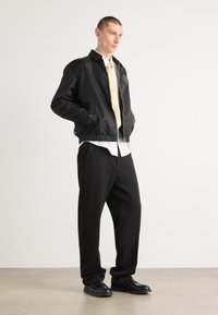 Veste bomber noire matelassée avec fermeture éclair, chemise blanche en dessous, cravate beige et pantalon noir à jambes larges. Des chaussures noires brillantes complètent le look.