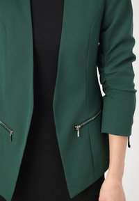 Zelený blazer vyrobený z hladkého materiálu s Přiléhavým střihem, který má zipové kapsy a na rukávech shromážděné detaily.