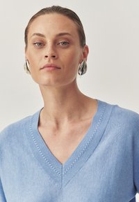 Pulover bleu deschis cu decolteu în V, având un guler cu nervuri și cusături decorative. Modelul poartă cercei metallici mari. Textură netedă.