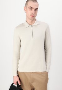Hele beežist värvi tõmblukuga pikad varrukad pullover kaelusega, valmistatud pehmest materjalist. Modell hoiab väikest musta kotti, jalas on taupe värvi püksid.