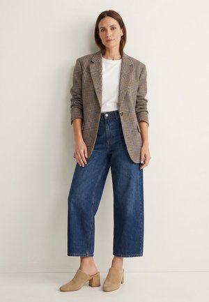 Frau steht an der Wand, trägt einen karierten Blazer, ein weißes Top, weite blaue Jeans und beige Mules mit Blockabsatz.