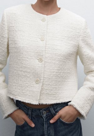 Femme portant une veste courte crème texturée avec des boutons sur un jean taille haute bleu foncé, mains dans les poches.