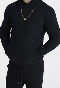 Felpa nera con cappuccio in tessuto morbido, con dettaglio a catena dorata e tasca frontale, abbinata a pantaloni neri coordinati.