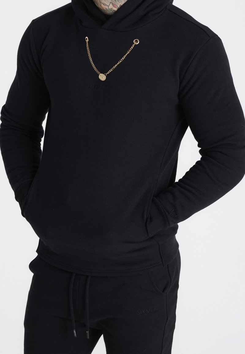 Felpa nera con cappuccio in tessuto morbido, con dettaglio a catena dorata e tasca frontale, abbinata a pantaloni neri coordinati.
