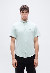 Νέος άνδρας με καστανά μαλλιά, φορώντας ανοιχτό πράσινο κοντομάνικο πουκάμισο με το λογότυπο "BOSS" και μαύρο παντελόνι, στέκεται μπροστά σε λευκό φόντο.