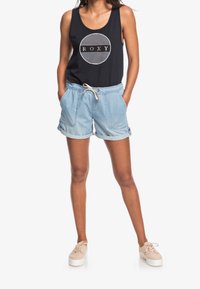 Zwart tanktop met een ronde logo, lichte denim shorts met opgerolde zomen en beige vetersneakers, gedragen door een model met handen in de zakken.