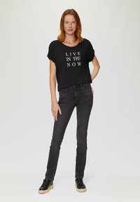 Zwarte T-shirt met korte mouwen en witte tekst "LEEF IN HET NU," gecombineerd met zwarte spijkerbroek en zwarte sneakers met een gestructureerde zool.