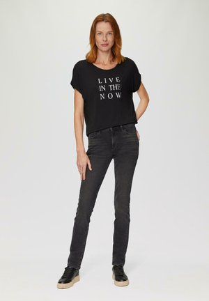 T-shirt noir à manches courtes avec le texte blanc "VIVRE DANS L'INSTANT", porté avec un jean noir et des baskets noires à semelle texturée.