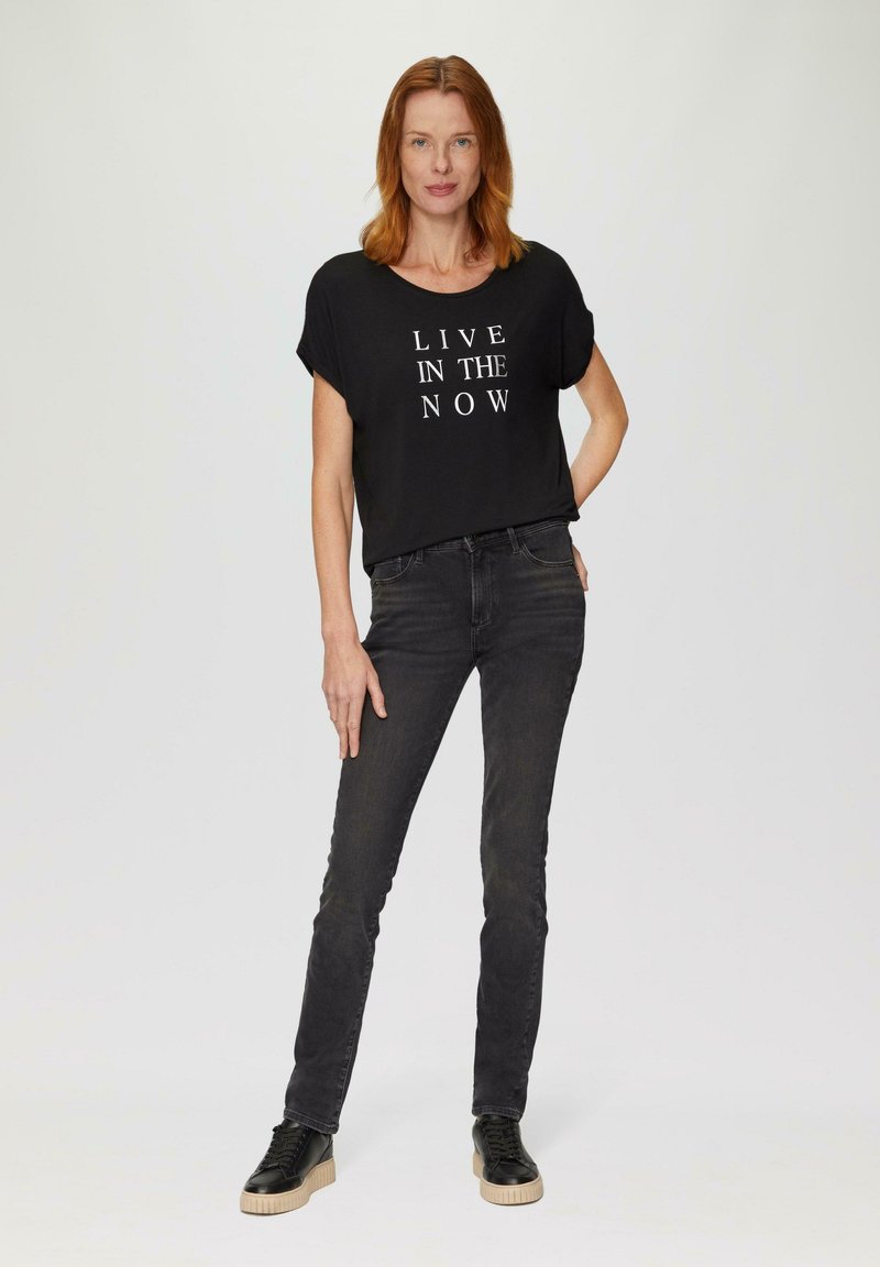 Zwarte T-shirt met korte mouwen en witte tekst "LEEF IN HET NU," gecombineerd met zwarte spijkerbroek en zwarte sneakers met een gestructureerde zool.