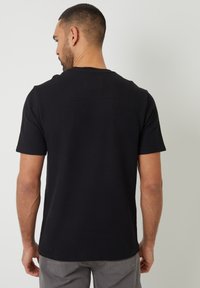 Schwarzes Kurzarmhemd mit strukturiertem Strickmuster, rundem Ausschnitt und dezentem Rückenschild-Detail. Getragen mit grauen Hosen.