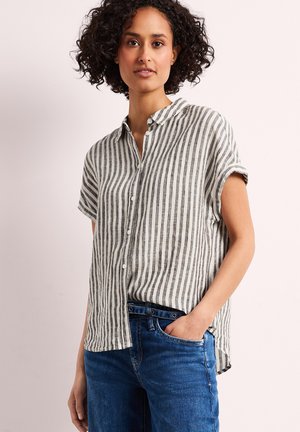 Femme aux cheveux bouclés portant une chemise à manches courtes rayée verticalement gris et blanc, boutonnée, et un jean bleu, main dans la poche.