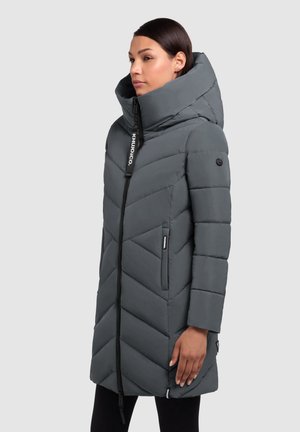 Femme portant un long manteau d'hiver matelassé gris charbon avec un col haut et une capuche, se tenant de profil devant un fond uni.