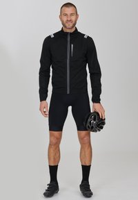 Svart cykeljacka med reflexdetaljer, bärs över svarta shorts och strumpor. Modellen håller en svart cykelhjälm.