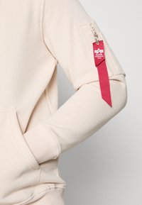 Felpa di un beige chiaro con polsi a coste, tasca frontale a marsupio e tasca con zip sulla manica sinistra con un'etichetta rossa con dettagli del logo.