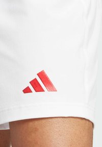 Shorts de sport blancs avec une finition texturée, présentant un logo Adidas rouge brodé proéminent sur le côté inférieur.