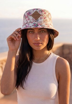 Jonge vrouw met lang donker haar die een bloemenprint bucket hat en een wit mouwloos topje draagt, terwijl ze de rand van de hoed aanraakt, buiten bij zonsondergang.